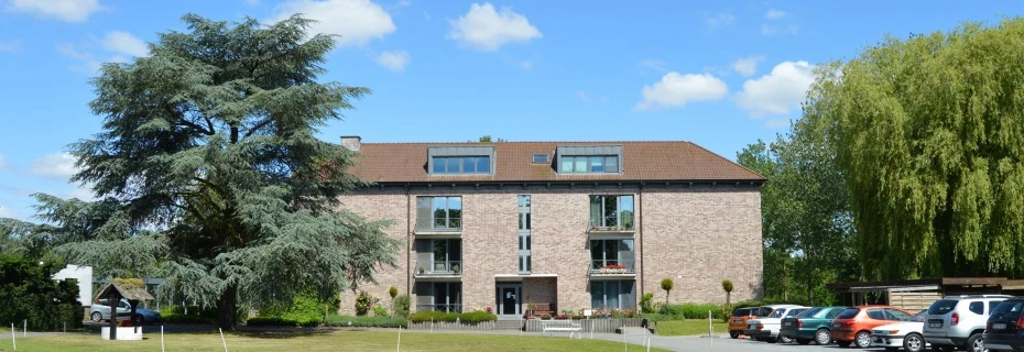 Bakstenen gebouw van vier verdiepingen in een park met grote coniferen en wilgen