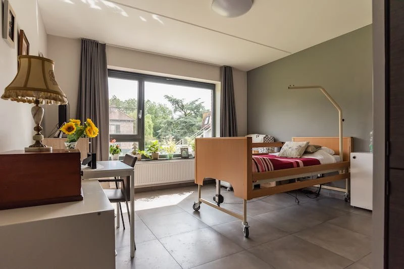Zorgkamer met bed, nachtkastje en bureau bij het raam op het groen